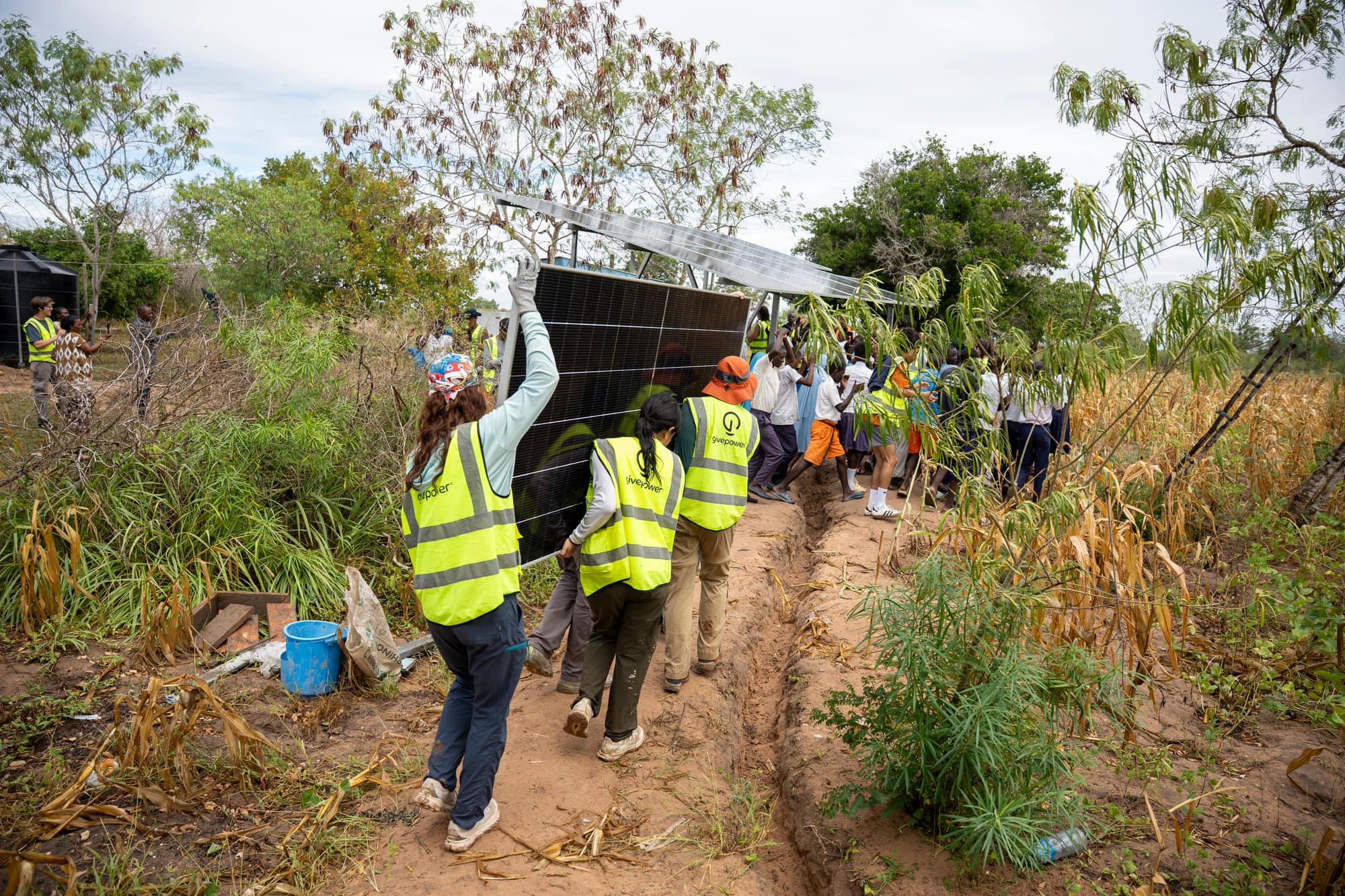 Volunteers in GivePower vests carrying a solar panel along a freshly dug trench to an install site